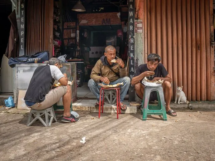 Three men eating near a store, with a cat in the city sitting beside them, captured by Marcel Heijnen.