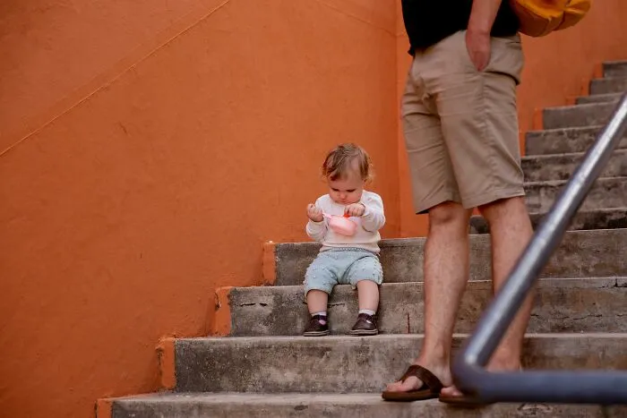 A child sitting on stairs next to an adult, holding a toy, with an orange wall in the background, conveying innocence and curiosity.