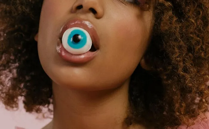 A person with curly hair holding a blue and white eyeball candy in their mouth, capturing a creepy moment.