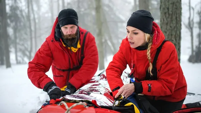 Two people in red jackets tending to a foil-wrapped stretcher in a snowy forest, representing traumatizing experiences.
