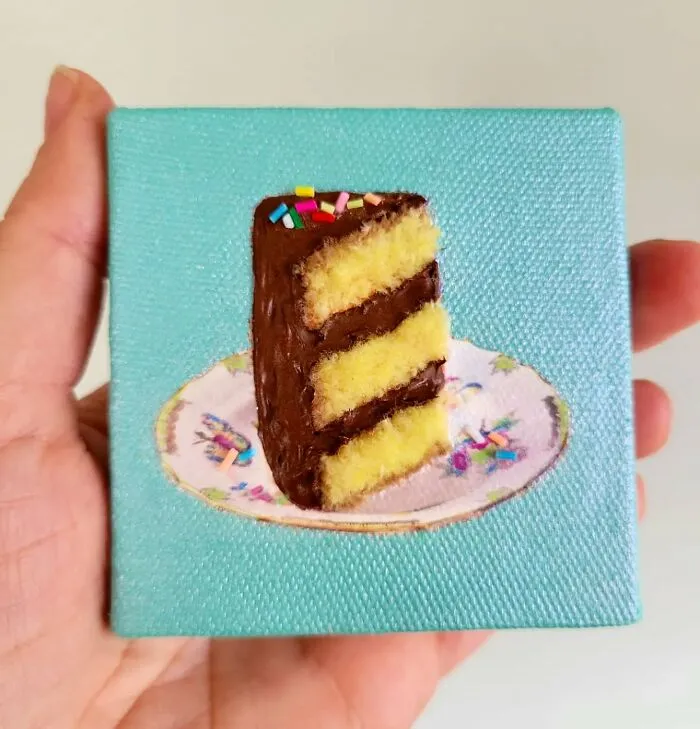 Hand holding a hyper-realistic embroidered dessert of a slice of chocolate cake on a small plate.