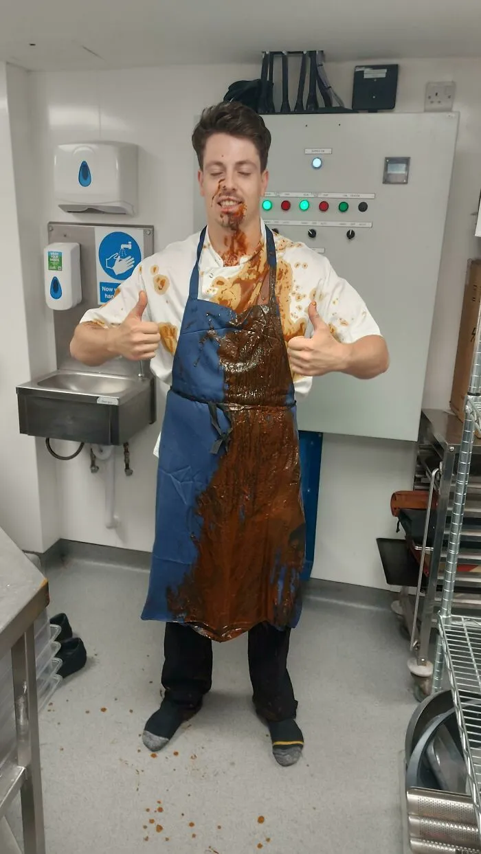 Worker in the food service industry with sauce all over their apron, giving thumbs up, in a kitchen setting.