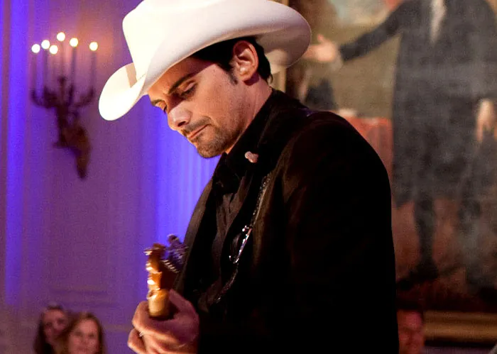Musician in a white cowboy hat playing guitar, performing on stage with a portrait in the background.