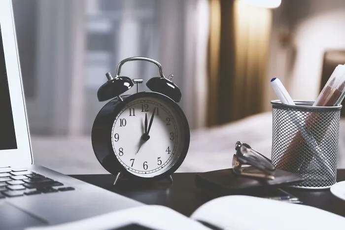 Vintage office desk setup with a classic alarm clock, open laptop, and pen holder, evoking humor about office routines.