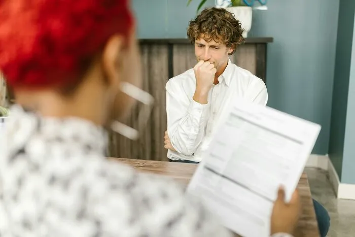 Person reviewing a document in an office, another looks thoughtful, capturing a humorous office moment.