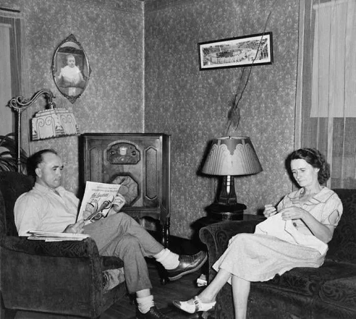 Personas en una sala de estar histórica, él lee un periódico y ella hace labores, junto a una antigua radio.