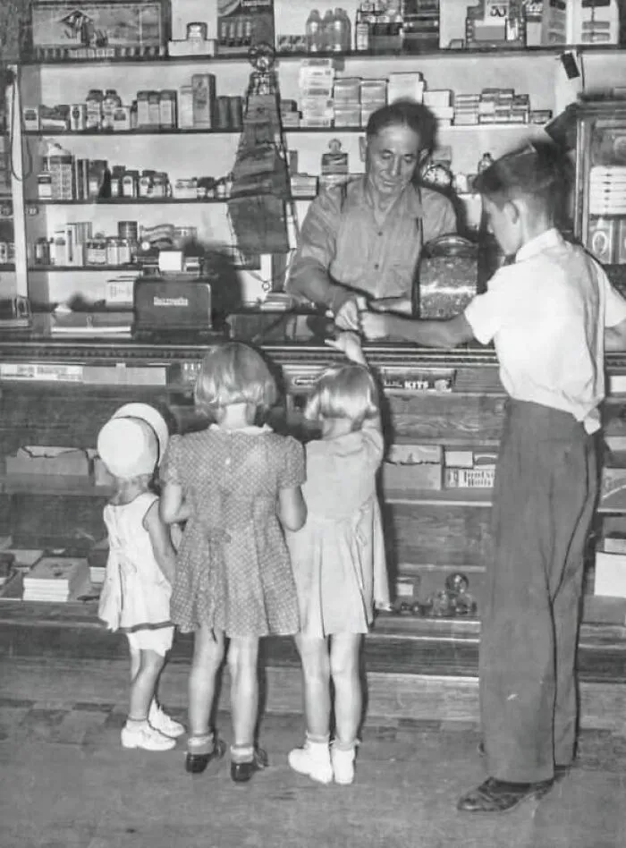 Niños comprando en una tienda antigua, atendidos por un tendero detrás del mostrador, evocando días pasados.
