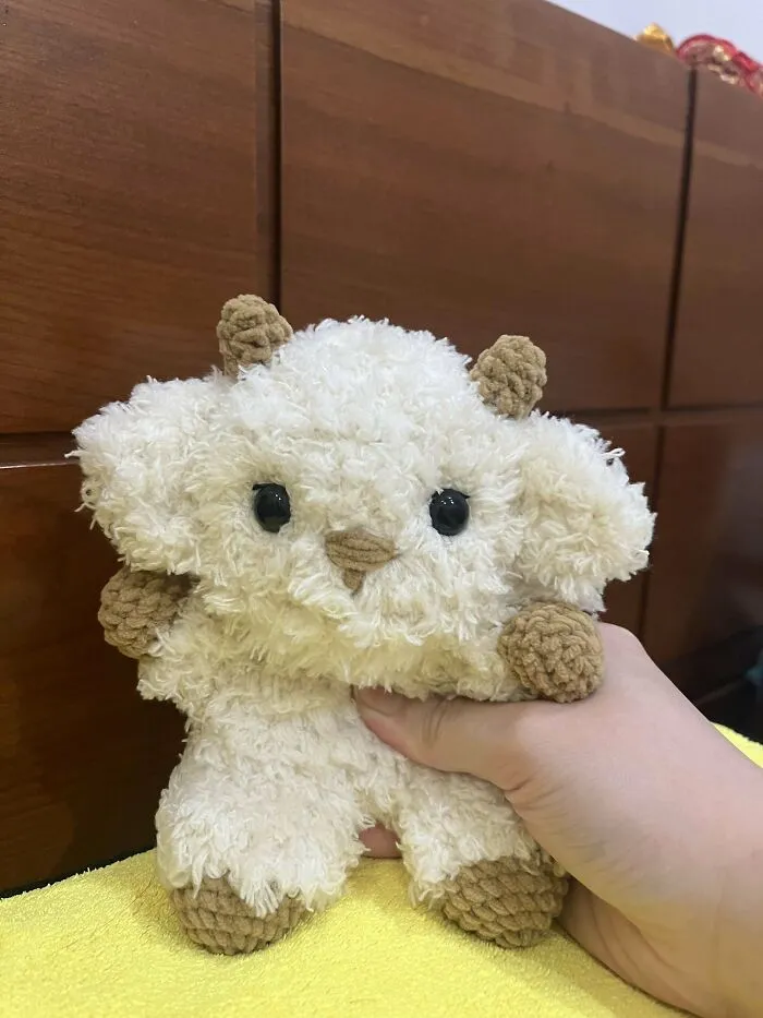 Hand holding a small, fluffy crochet animal with black eyes and brown details, showcasing incredible crochet craftsmanship.