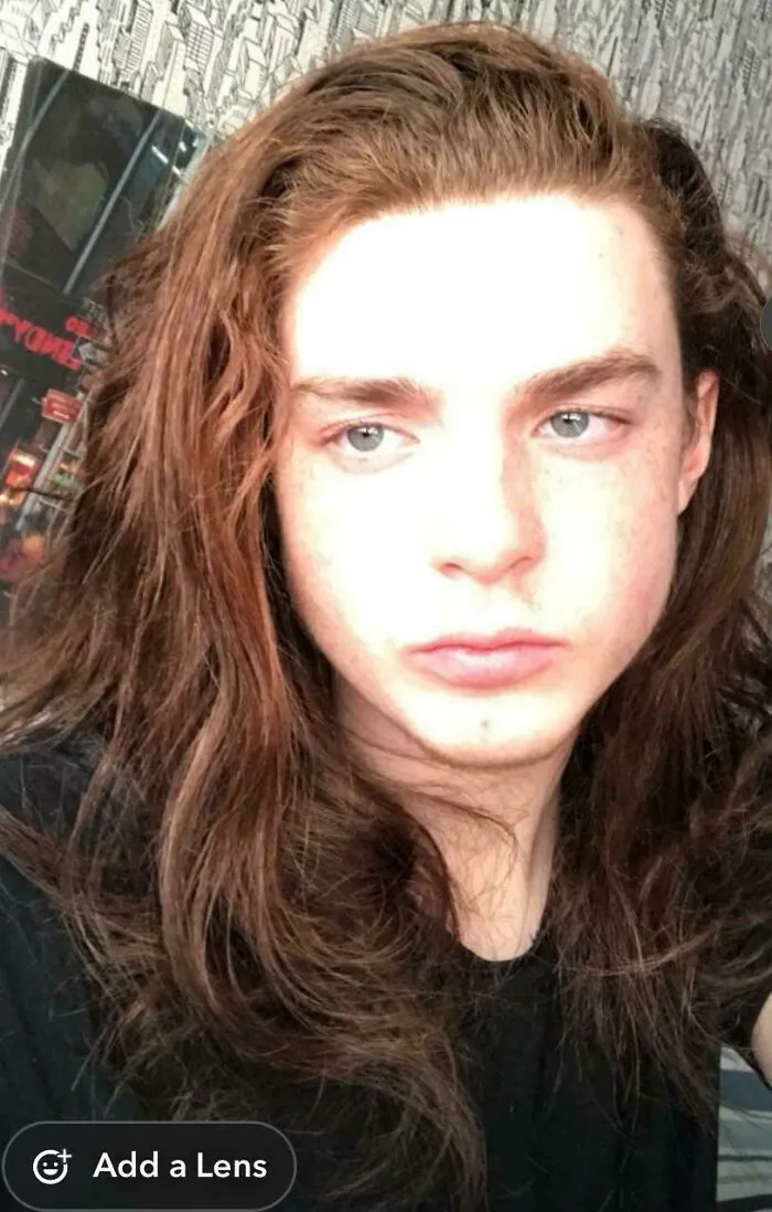 Young man with fierceflow beautiful hair posing indoors with long brown wavy hair and a neutral expression.