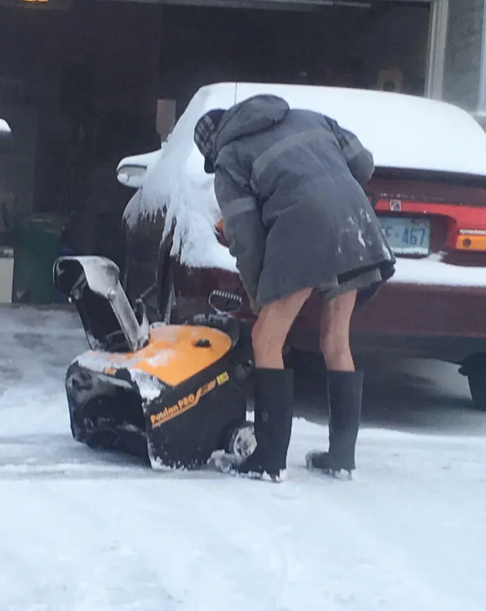 Persona en Canadá usando soplador de nieve, vistiendo abrigo largo, botas y gorro en una mañana nevada.