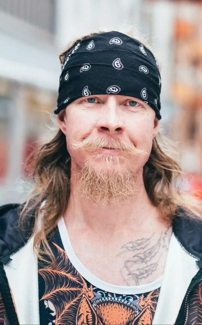 Long-haired man with a bandana, featuring a unique mustache and tattoo, as part of street portraits by a photographer.