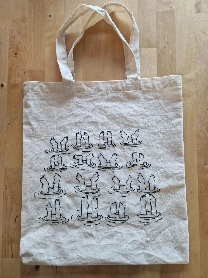 Beige tote bag featuring embroidery of various small feet and legs poking out of water in black thread.