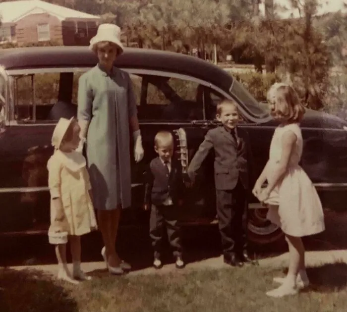 Vintage family photo with children and woman dressed in mid-century attire standing by classic car, candid glimpses into past.