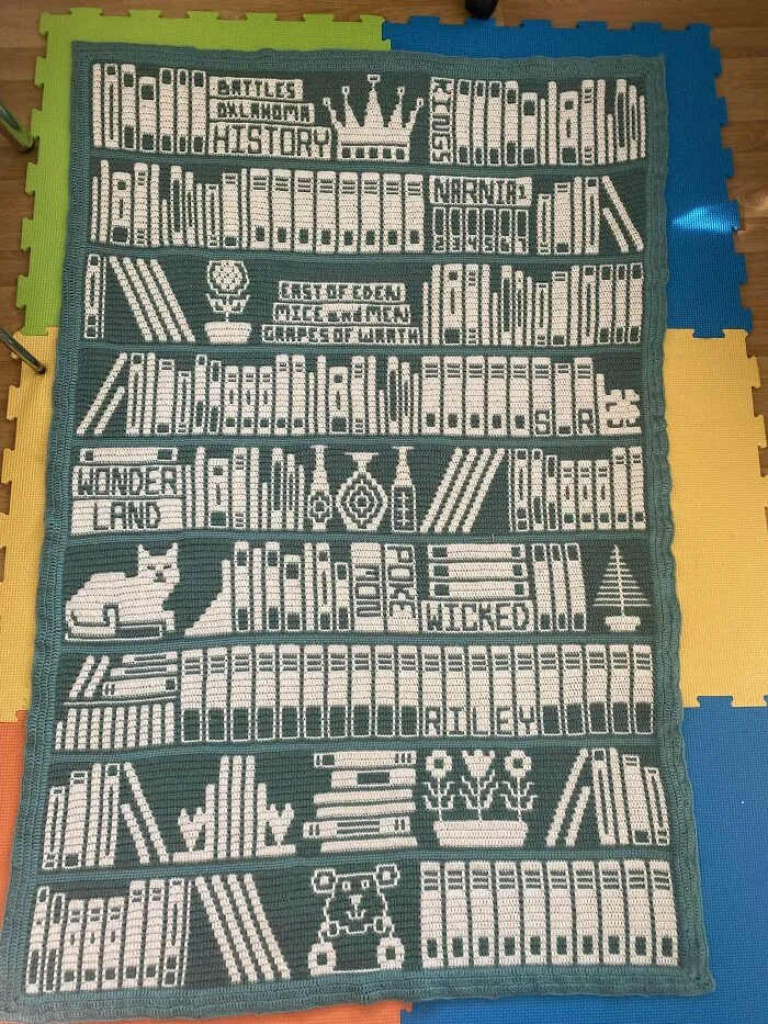 Crochet blanket designed to look like bookshelves filled with books, flowers, and decorative items, showcasing intricate crochet skills.
