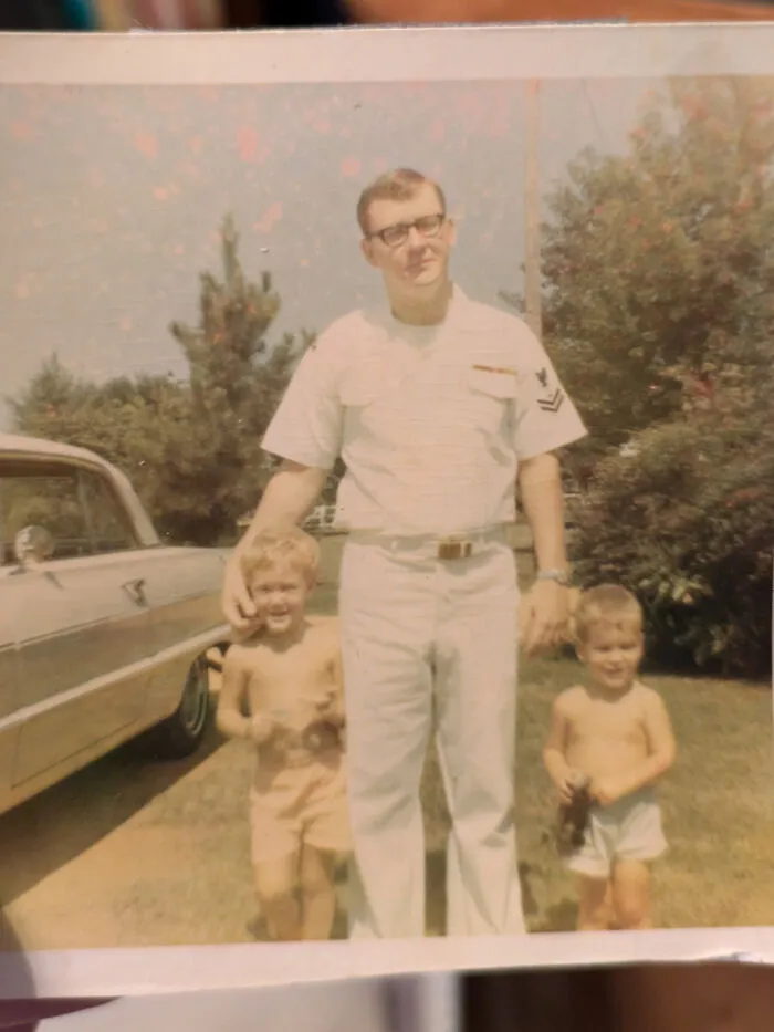 Vintage candid glimpse of a man in uniform with two young children outdoors next to a classic car in summer.