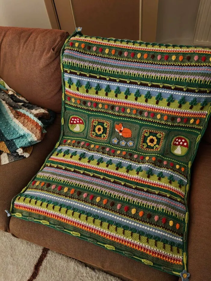 Colorful crochet blanket with nature-inspired patterns including mushrooms, fox, and flowers on a brown couch, crochet enthusiasts creation.