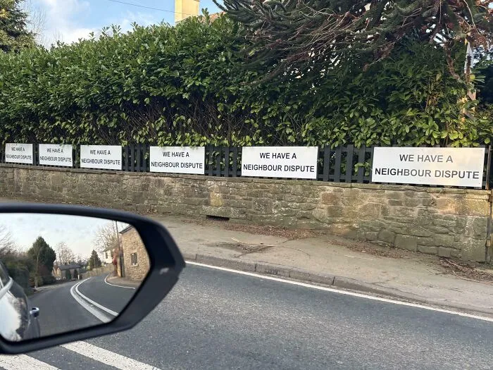 Carteles pasivo-agresivos en una valla anuncian disputa vecinal, vistos desde un coche en movimiento.