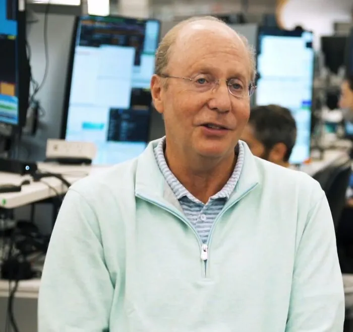 Man in light sweater speaking in an office with multiple monitors, representing the richest people in the world wealth comparison.