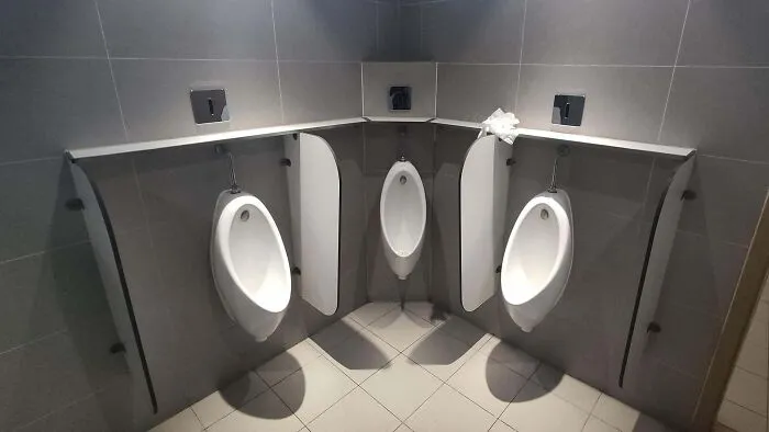 Urinales en esquina mal diseñados en un baño público, mostrando problemas de privacidad. Diseño curioso y poco funcional.