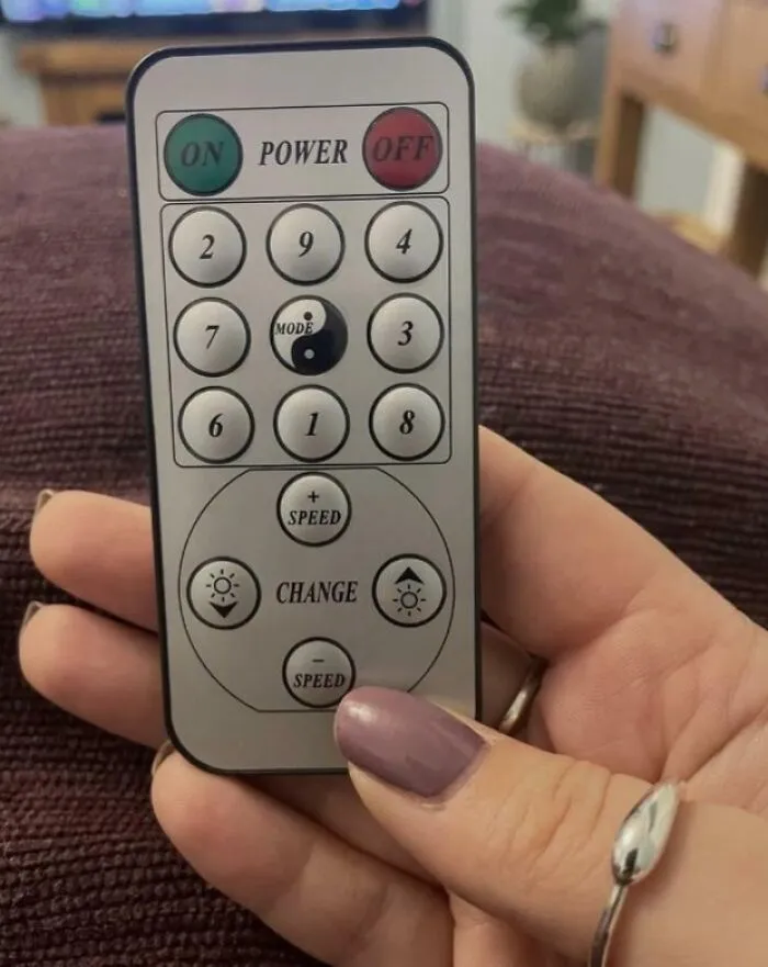 Control remoto con botones confusos, discretamente sostenido entre dedos con uñas pintadas, bajo una luz suave.