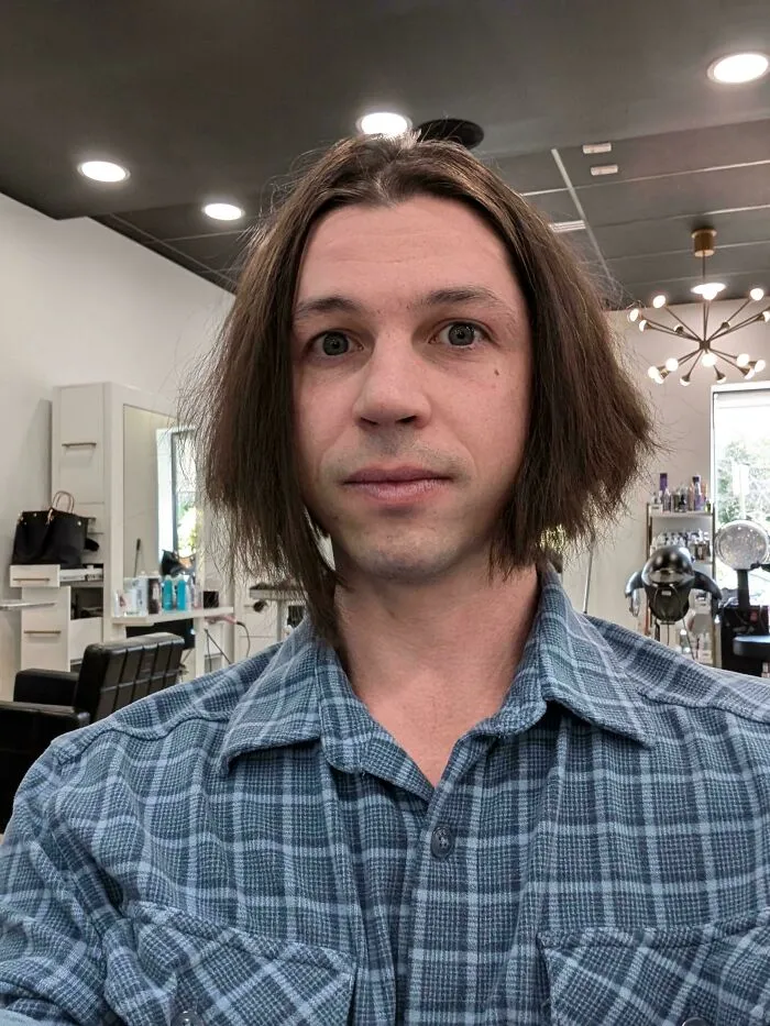Man with an unfortunate haircut, long shaggy brown hair, looking at the camera in a salon.