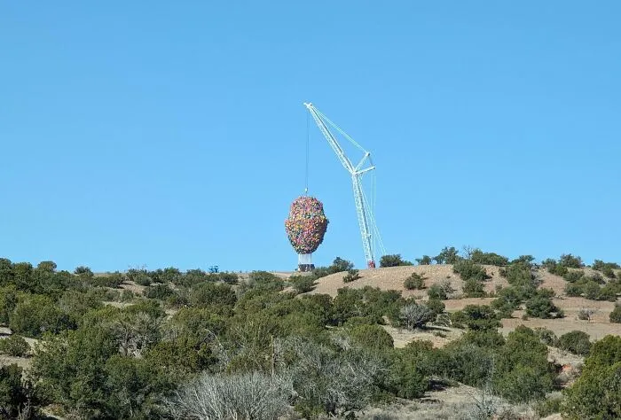 Grúa levantando una gran escultura en forma de cabeza hecha de globos, rodeada de paisaje natural.