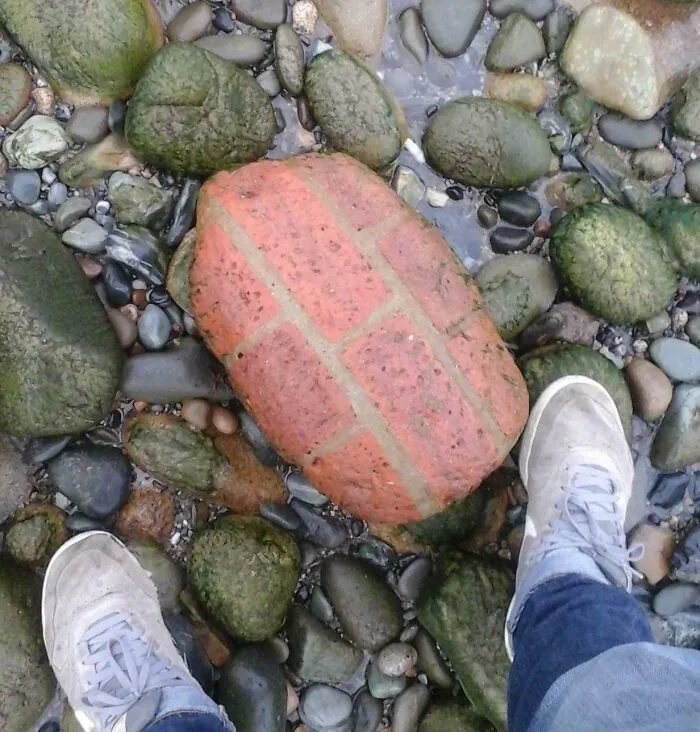 Weathered brick among pebbles, showing effects of time and use, with two sneaker-clad feet visible.