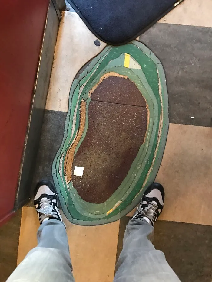 Worn-out floor mat showing layers, transformed over time and use, viewed from above with a person's shoes visible.