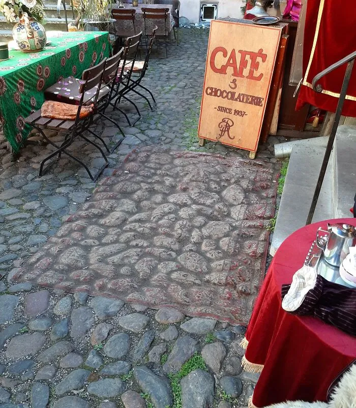 Café sign and worn rug on cobblestone path, showing effects of time and use.