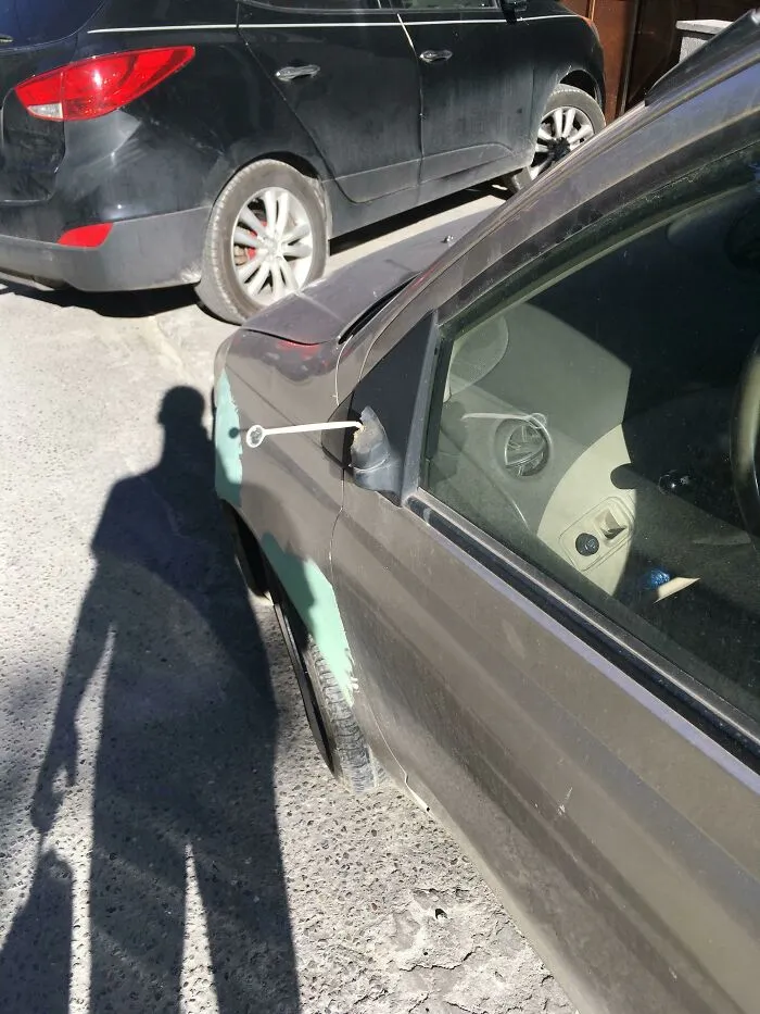 Espejo lateral del coche mal reparado con cinta adhesiva improvisada y sombra en el suelo.