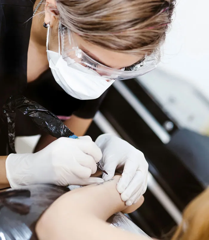 Tattoo artist with safety gear working on a client's arm in a tattoo studio.