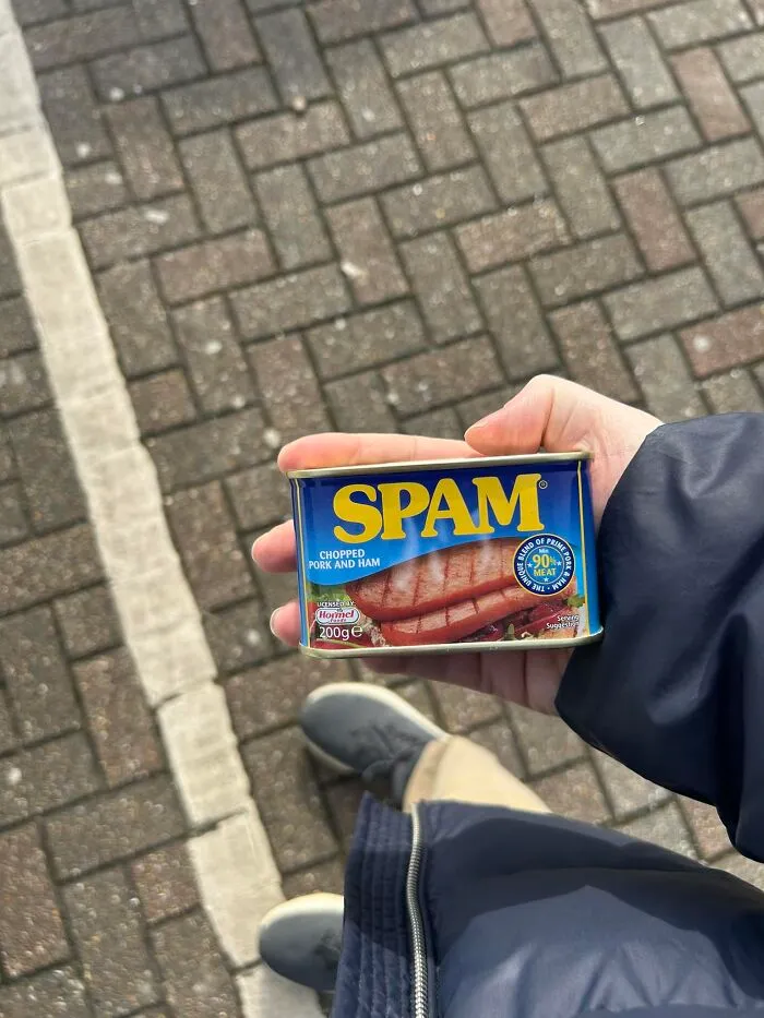 Person holding a can of Spam on a pavement, highlighting British humor.