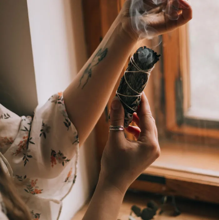 Person with tattoo on arm holding a smudge stick by the window.
