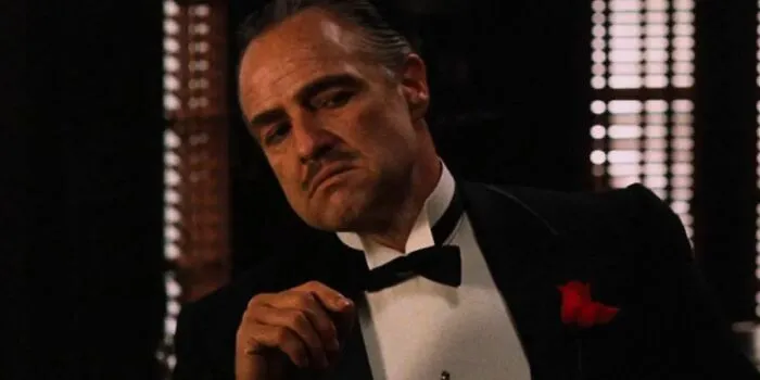 Man in a tuxedo with a rose, lit by window blinds, embodies hyped expectations.
