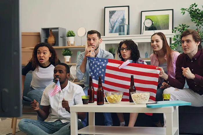 Group of friends watching TV, holding USA flag, with snacks and drinks on the table, showcasing normal activities in America.