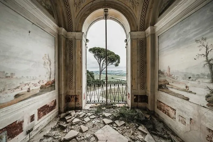 Abandoned place with crumbling floor and mural walls, overlooking a lush landscape through an arched window.