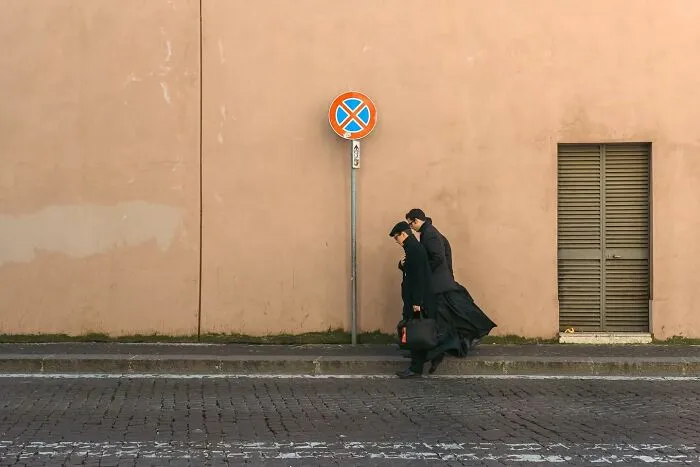Two people in black coats walk past a no parking sign on a cobblestone street, showcasing street photography by Andrea Torrei.