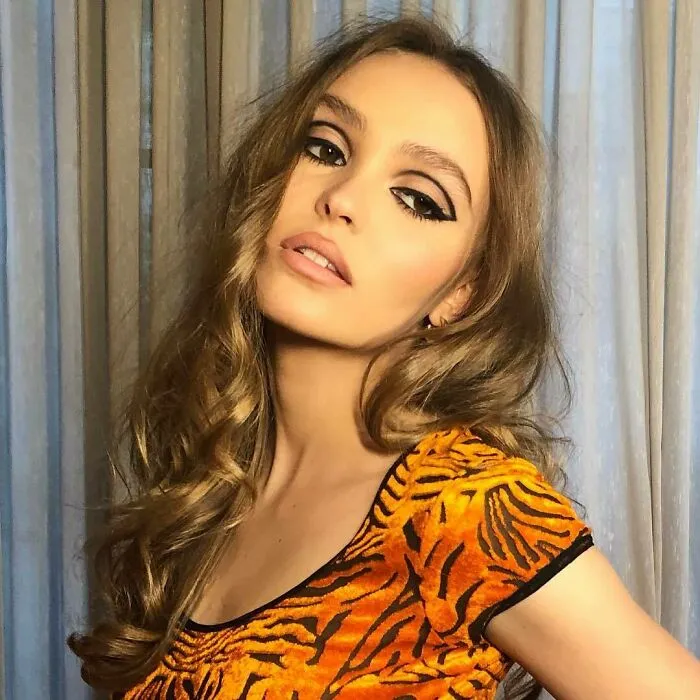 A woman in a patterned orange top poses against curtains, embodying not-self-made-celebs style.