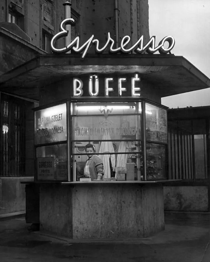 Historical pic of an espresso stand with a vendor inside, featuring vintage neon signs and nostalgic urban backdrop.