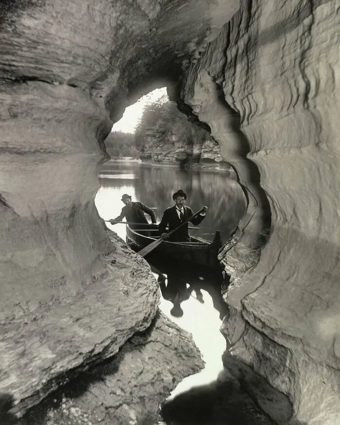Two men in a vintage boat navigate a narrow canyon, evoking historical nostalgia with scenic reflections on the water.