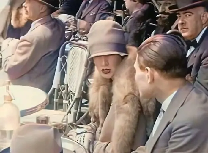 Elegant 1920s couple at an outdoor gathering, surrounded by spectators, evokes nostalgia with vintage attire and hats.