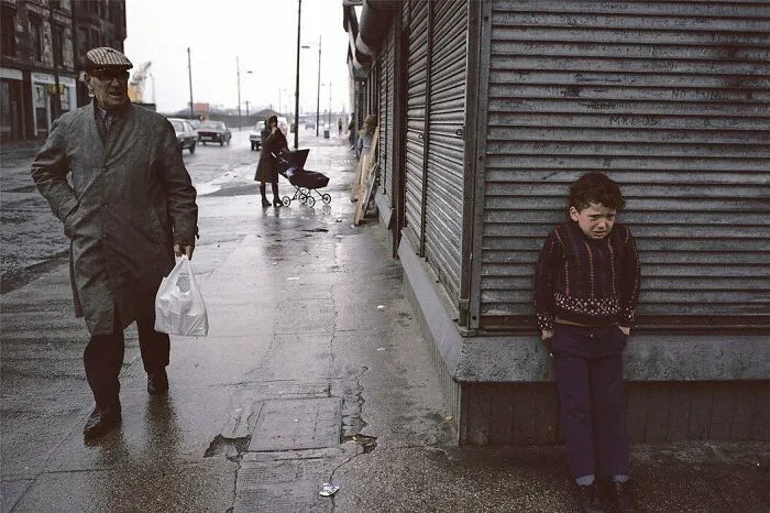 Man with bag walks past a boy on a rainy street, evoking historical nostalgia.