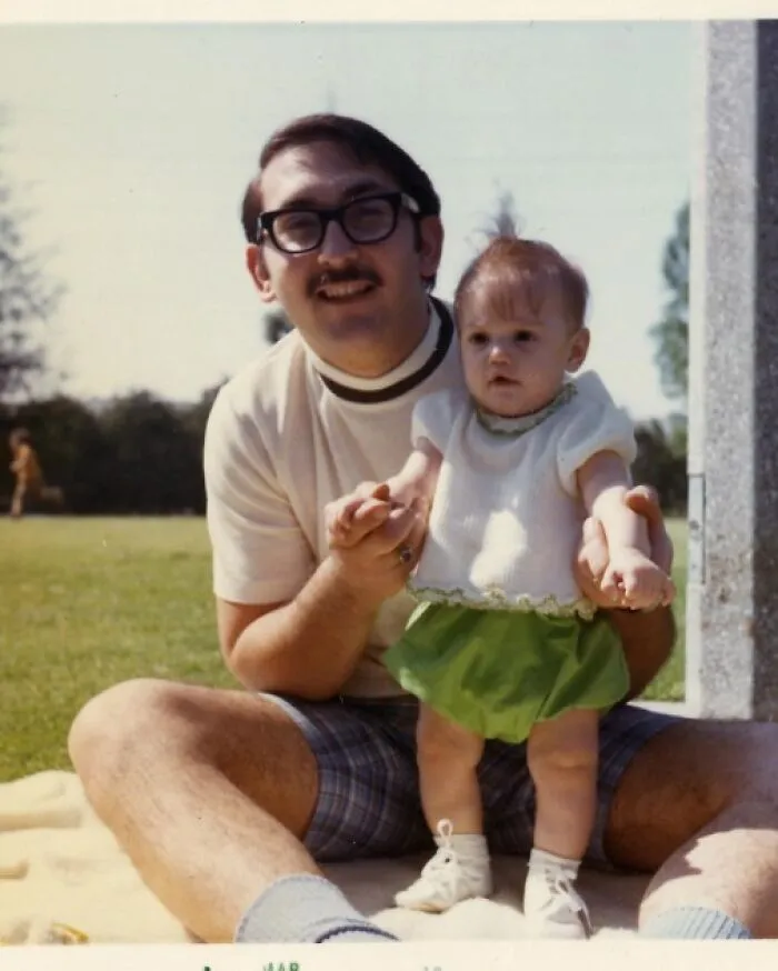 A man with glasses holds a baby in a park, capturing a moment from the past.