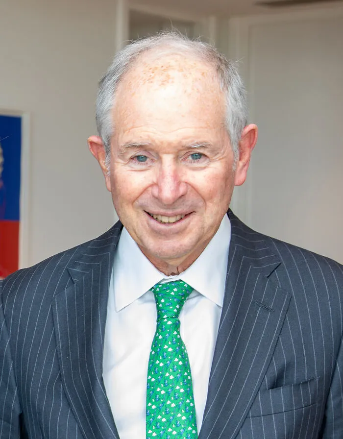 Elderly man in pinstripe suit and green tie smiling indoors, representing the richest people in the world with vast wealth.