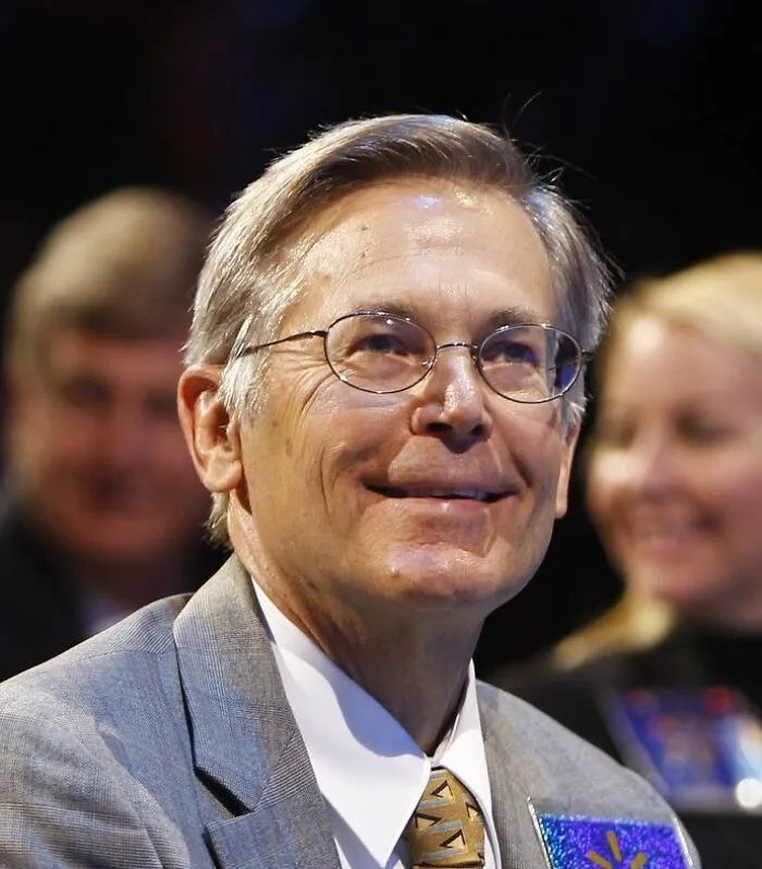 Older man wearing glasses and a suit, smiling at an event highlighting the richest people in the world wealth.