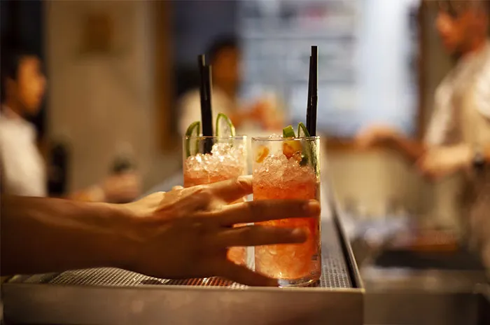 Hand reaching for a cocktail on a bar counter, with a bartender in the background.