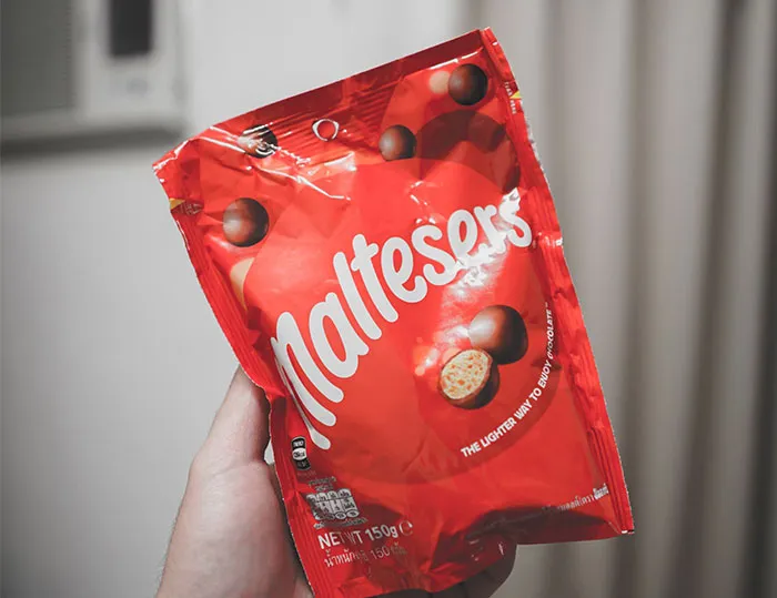 A hand holding a red bag of Maltesers chocolates indoors.