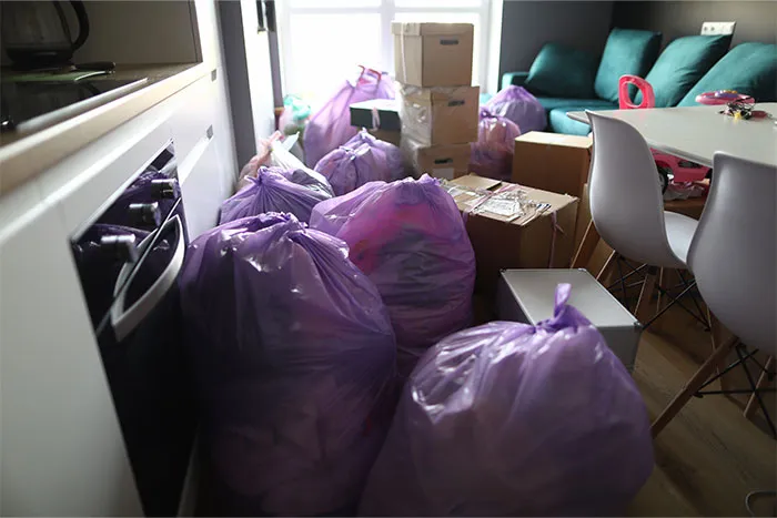 Purple trash bags and cardboard boxes fill a room with teal pillows and white chairs, hinting at a cluttered domestic scene.
