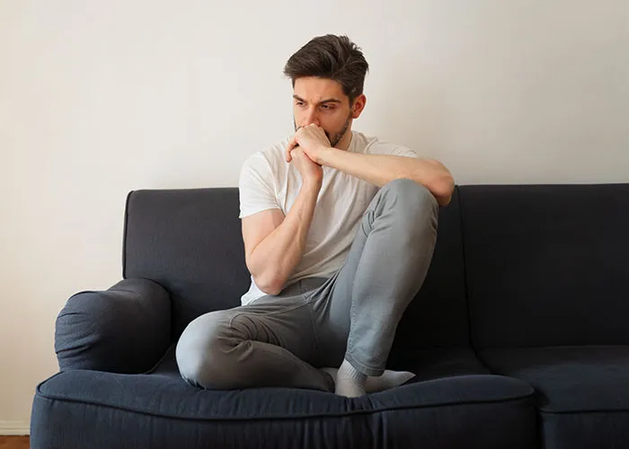 Young man in deep thought sitting on a couch in a casual home setting, reflecting on questions about men.