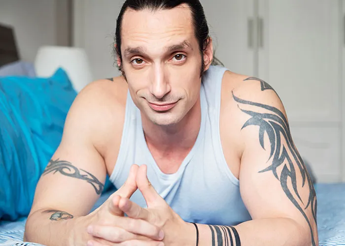 Man with tattoos lying on a bed, wearing a light blue tank top, and looking at the camera.
